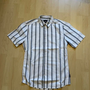 Randig kortärmad skjorta - Snygg kortärmad skjorta från Only & Sons med vertikala svarta och grå ränder på vit botten. Klassisk krage och knäppning framtill. Perfekt för en avslappnad och stilren look.