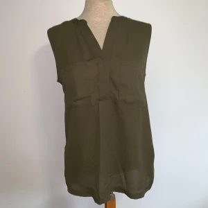 Olivgrön ärmlös blus från H&M - Snygg olivgrön blus från H&M i storlek S. Modellen är ärmlös med v-ringad hals och har två dekorativa bröstfickor framtill. Materialet är lätt och skönt, perfekt för varmare dagar. 