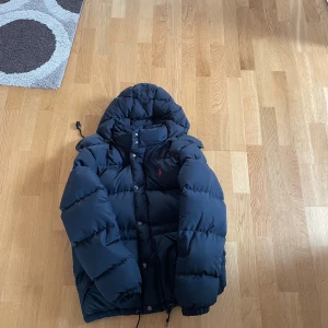 Mörkblå pufferjacka från Polo Ralph Lauren - Säljer en mörkblå pufferjacka från Polo Ralph Lauren med huva och röd broderad logga på bröstet. Jackan har dragkedja och knappar framtill samt snörning i nederkant. Perfekt för kalla dagar och riktigt snygg streetstil.