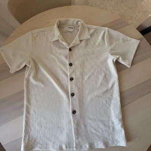 Vit kortärmad skjorta från Selected - Snygg vit skjorta från Selected med diskreta tunna ränder och klassisk krage. Skjortan har korta ärmar och mörka knappar framtill. Perfekt för en clean och stilren look.