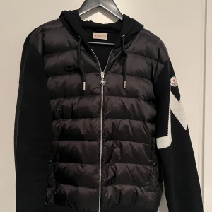 Svart Moncler pufferjacka med mönster - Svart pufferjacka från Moncler med stickade ärmar och huva. Jackan har dragkedja framtill, snörning vid huvan och ett snyggt zigzag-mönster i vitt på ena ärmen. Materialmix av polyester och stickad textil ger en cool kontrast. Perfekt för dig som gillar streetstyle.