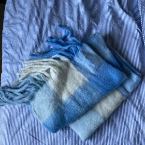 Blåvit rutig ullhalsduk med fransar - Mysig och fluffig halsduk i rutigt mönster med olika nyanser av blått och vitt. Den har långa fransar i ändarna och är tillverkad i mjuk ull som känns skön mot huden. Perfekt att svepa runt halsen för extra värme under kyliga dagar.