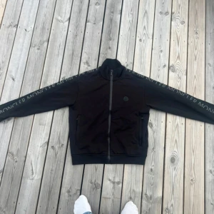 Svart Moncler zip up kofta - DEN ÄR ÄKTA! Pris kan diskuteras vid snabb affär!! Använd fåtal gånger!! Svart kofta från Moncler med dragkedja framtill och Moncler-logga på bröstet. Snygga detaljer med Moncler-tryck längs ärmarna och två fickor med dragkedja. Koftan har hög krage och är tillverkad i mjukt bomullsmaterial.