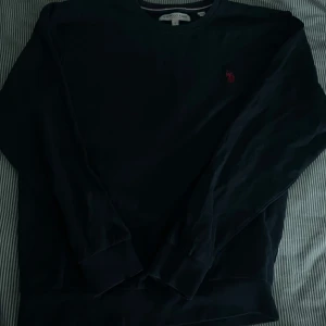 Svart sweatshirt från U.S. Polo Assn. - Svart sweatshirt från U.S. Polo Assn. med klassisk rund hals och broderad röd logga på bröstet. Tröjan har långa ärmar och ribbade muddar vid ärmslut och nederkant. Perfekt för en chill och stilren look.