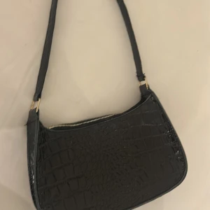 Svart axelväska i croco-imitation - Snygg svart axelväska med croco-mönstrad yta och guldfärgade detaljer. Väskan har en smidig dragkedja upptill och en stilren, avlång form som passar perfekt till din outfit. Materialet är glansigt syntet och ger en lyxig känsla.