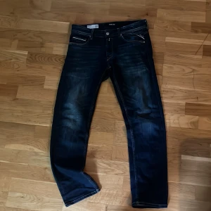 Replay Grover jeans straight fit blå - Snygga Replay Grover jeans i mörkblå tvätt med raka ben och klassiska kontrastsömmar. Jeansen har fem fickor, läderpatch bak och coola slitningar framtill. Perfekta för dig som gillar en clean och tidlös look. Storlek 31/30