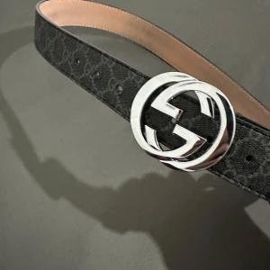 Gucci bälte  - Snyggt svart bälte från Gucci med klassiskt GG-mönster och ett stort, rundat spänne i silver. Bältet är gjort i skinn och har en lyxig känsla med beige insida. Perfekt accessoar för att lyfta vilken outfit som helst.