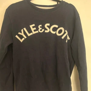 Mörkblå Lyle & Scott sweatshirt - Mörkblå sweatshirt från Lyle & Scott med stort vitt tryck på bröstet. Klassisk rund halsringning och långa ärmar. Tröjan är i mjuk bomull och passar perfekt för en chill och stilren look.