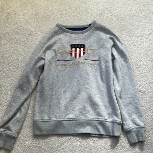 Grå sweatshirt från GANT med brodyr - Säljer en grå sweatshirt från GANT med broderad logga och sköld på bröstet. Tröjan har rund halsringning, långa ärmar och ribbade muddar. Mjuk insida som känns riktigt skön mot huden. Perfekt för dig som gillar klassisk och sportig stil.