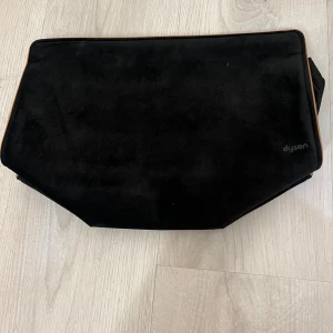 Svart kuvertväska från Dyson i mocka - Stilren svart kuvertväska från Dyson i mjuk mocka med bruna kanter. Väskan har en minimalistisk design och diskret logga i hörnet. Perfekt för att bära småsaker och accessoarer, och ger en clean look till outfiten.
