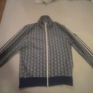 Mönstrad track jacket från Lacoste M - Säljer en blå och grå track jacket från Lacoste i storlek M. Jackan har ett grafiskt mönster, dragkedja framtill och vita ränder längs ärmarna. Klassisk Lacoste-logga på bröstet. Tillverkad i polyester och bomull, perfekt för en sportig och trendig look. Skickar samma dag eller dagen där på