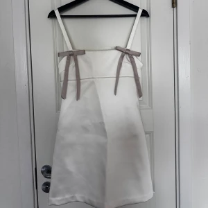 Vit kort klänning med rosetter - Supersöt vit kort klänning från Zara i satinliknande material. Klänningen har smala axelband och två ljusgrå rosetter framtill som ger en gullig touch. Stängs med dragkedja i ryggen.