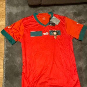 Röd Marocko fotbollströja Puma - Säljer en officiell Marocko fotbollströja från Puma i rött med gröna detaljer vid krage och ärmslut. Tröjan har korta ärmar, v-ringad hals och emblem på bröstet. Materialet är lätt och andas, perfekt för match eller träning.