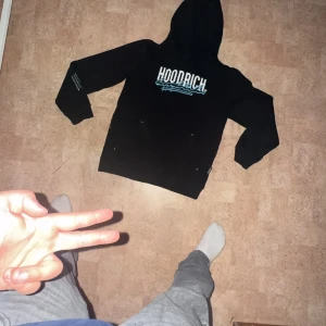 Svart HOODRICH hoodie med tryck - Svart hoodie från HOODRICH med vit och blå logga framtill och textdetaljer på ärm och rygg. Klassisk huva och känguruficka. Snygg streetwear-stil, perfekt för dig som gillar en chill och trendig look.