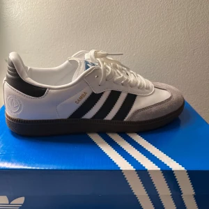 Adidas Samba OG vita och svarta sneakers - Adidas Samba OG sneakers i vitt och svart med klassiska tre ränder på sidan, brun gummisula och detaljer i mocka framtill. Skorna har snörning och låg profil, samt guldfärgad Samba-text på sidan. Perfekta för dig som gillar retrostil och streetwear.