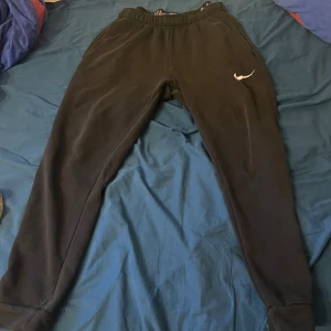 Svarta Nike Dri-FIT mjukisbyxor L - Svarta mjukisbyxor från Nike med Dri-FIT-teknologi och klassisk Swoosh-logga på benet. Byxorna har resår i midjan, vanlig passform och muddar vid bensluten. Perfekta för chill eller träning, tillverkade i ett mjukt och funktionellt material.