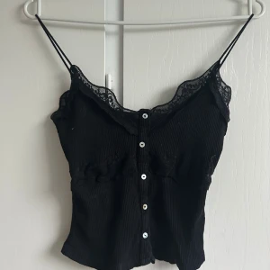 Svart ribbad linnetopp med spets - Supersnygg svart linnetopp med tunna axelband, ribbad struktur och spetsdetaljer längs urringningen. Knappar framtill ger en extra touch. Perfekt att styla med jeans eller kjol för en trendig look.