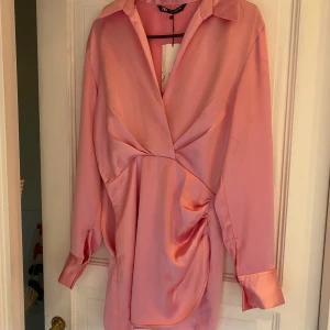 Rosa omlottklänning från Zara - Säljer en superfin rosa kortklänning från Zara i glansigt satinmaterial. Klänningen har omlottdesign, v-ringning och långa ärmar med breda manschetter. Perfekt för dig som vill sticka ut med en cool och trendig look.