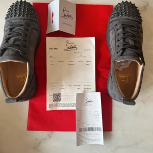 Grå Christian Louboutin sneakers med nitar - Snygga grå sneakers från Christian Louboutin i mocka med ikoniska röda sulor och svarta nitar på tån. Skorna har rund tå, snörning och platt sula. Perfekta för dig som vill sticka ut med en lyxig och edgy stil.!!kvitto ingår!!