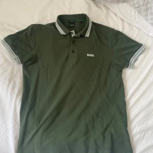 Grön BOSS pikétröja med vit logga - Snygg grön pikétröja från BOSS med vit broderad logga på bröstet. Klassisk krage med knappar och vita detaljer på krage och ärmslut. Tillverkad i bomull för skön känsla och bra passform. Perfekt för dig som gillar stilrena och sportiga plagg. Kvittot finns