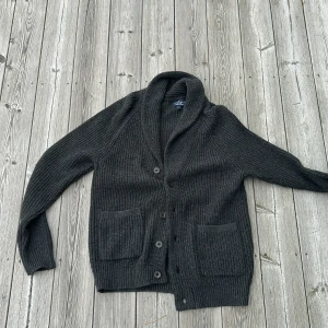 Mörkgrå kofta från Hackett i merinoull - Säljer en mörkgrå, ribbstickad kofta från Hackett London i lyxig mix av merinoull och kashmir. Koftan har sjalkrage, knäppning med fyra knappar och två stora fickor framtill. Perfekt för lager-på-lager och chill höst- och vinterstil.