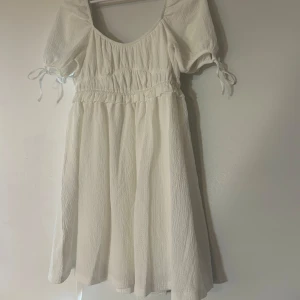 Vit puffärm klänning från Gina Tricot - Supersöt vit kortklänning från Gina Tricot med puffärmar och knytband vid ärmslut. Klänningen har en omlottliknande midjedetalj.