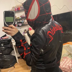 Svart och röd Spiderverse hoodie - Cool svart hoodie inspirerad av Spiderverse med röda spindelnätsdetaljer och stora vita ögon på huvan. Röd text på ryggen och röda detaljer längs ärmarna. Perfekt för dig som vill sticka ut och gillar superhjältar.
