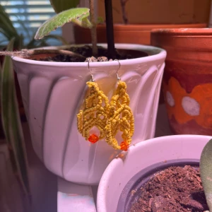 Handgjorda gula makraméörhängen - Unika örhängen i makramé med knallgul tråd och en orange pärla längst ner. De har en snygg, bohemisk form och är perfekta för dig som vill sticka ut med färg och kreativ stil. Krokar i metall för enkel användning.