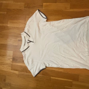 Vit pikétröja från Massimo Dutti - Vit pikétröja från Massimo Dutti med klassisk krage och svarta detaljer på krage och ärmslut. Casual fit och tillverkad i bomull, perfekt för en clean och stilren look. Kortärmad modell som funkar året runt.