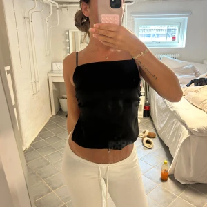 Svart sidentopp från Zara med öppen rygg  - Säljer en svart linnetopp från Zara med tunna axelband och snygg omlottdetalj i ryggen som knyts till en rosett. Toppen har rak halsringning och är croppad, perfekt för en trendig look. Materialet är mjukt och har en lätt glansig finish.