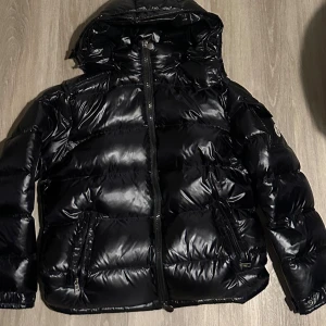 Svart Moncler dunjacka med huva - Säljer en svart, glansig dunjacka från Moncler med huva och dragkedja framtill. Jackan har Moncler-logga på ärmen, två fickor med dragkedja och quiltad design. Perfekt för kalla dagar när du vill hålla stilen och värmen. Du kan lägga telefonen på märket på sidan av jackan så kommer du komma in på Monclers hemsida 