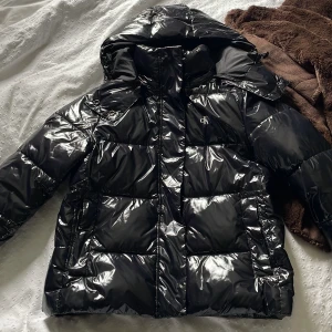 Svart pufferjacka Calvin Klein Jeans - Säljer en svart, glansig pufferjacka från Calvin Klein Jeans med huva och dragkedja. Jackan har logga på bröstet och ärmen, quiltad design och är perfekt för kalla dagar. Oversized känsla och riktigt snygg streetstil.