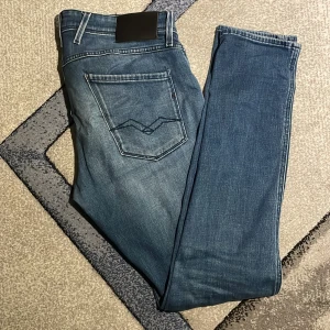 Replay Anbass blå jeans slim fit - Säljer ett par snygga Replay Anbass jeans i blå tvätt med slitningar och klassiska detaljer. Jeansen har slim fit-passform, fem fickor och tydliga Replay-loggor. Perfekta för dig som gillar en modern och stilren look. 34W34L