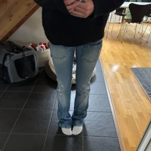 Blå bootcut jeans med slitningar - Säljer ett par blå bootcut jeans med snygga slitningar och stora fickor bak med knappar. Jeansen har låg midja och är i klassisk denim med ljus tvätt. Perfekta för dig som gillar en avslappnad och trendig look.