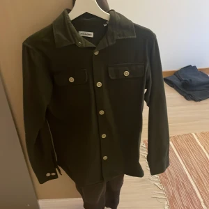 Mörkgrön overshirt från Jack & Jones XS - Stilren mörkgrön overshirt från Jack & Jones i storlek XS. Skjortan har klassisk krage, två bröstfickor med knappar och långa ärmar. Perfekt att bära öppen eller stängd för en avslappnad look.