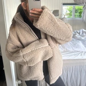Pile jacka  - Supermysig beige teddyjacka med oversized passform och lång ärm. Jackan har en hög krage och stängs med dragkedja framtill. Perfekt för kyliga dagar när du vill vara både varm och snygg. Passar till allt och är lätt att styla.