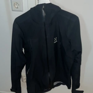 Svart Haglöfs Gore-Tex vindjacka - Svart vindjacka från Haglöfs i Gore-Tex-material. Jackan har huva, hel dragkedja framtill och två snedställda fickor med dragkedja. Perfekt för blåsiga och regniga dagar, stilren och funktionell design.