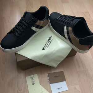 Burberry skor - Snygga svarta sneakers från Burberry med den ikoniska beige och svarta rutan på sidorna. Skorna har vita sulor, svarta snören och detaljer i skinn. Perfekta för dig som vill ha en stilren och lyxig look nu till hösten/vintern. Säljer de pga de inte passa mig! Pris kan diskuteras!!!