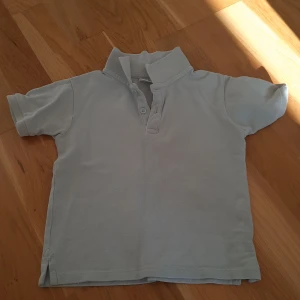 Ljusblå pikétröja från Zara, strl 98 - Ljusblå pikétröja från Zara i storlek 98, med klassisk krage och två knappar framtill. Tröjan har korta ärmar och är tillverkad i mjuk bomull