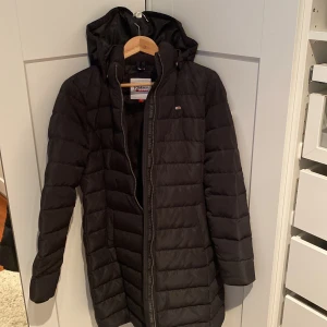 Svart lång dunjacka Tommy Jeans - Säljer en svart lång dunjacka från Tommy Jeans med huva och dragkedja. Jackan har quiltad design och logotyp på bröstet och ärmen. Perfekt för kalla dagar och har en stilren look som funkar till många outfits. nästan helt ny, har bara använt den 1 gång pågrund av att den var för liten.