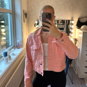 Croppad rosa jeansjacka från Gina Tricot - Snygg croppad jeansjacka i ljusrosa/skrikrosa från Gina Tricot. Jackan har klassisk krage, två bröstfickor med lock och silverfärgade knappar framtill. Tight modell. Perfekt att slänga över en t-shirt för en chill look. Långärmad och med skön passform.