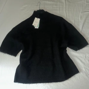 Svart stickad tröja med funnel neck L - Svart stickad tröja från Ginatricot i storlek L med funnel neck och korta ärmar. Tröjan har en mjuk och fluffig känsla, perfekt för lager-på-lager. Enkel och stilren design som funkar till många outfits. Aldrig använd, därav prislappen på! Hör av dig vid frågor 🥰