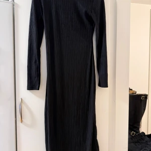 Svart ribbad långklänning XS - Svart långklänning från NA-KD i storlek XS. Klänningen har ett ribbat tyg, lång ärm och en snygg fyrkantig urringning. Materialet är mjuk och stretchig polyester med lite elastan för extra komfort. Perfekt för dig som gillar stilrena och minimalistiska looks.