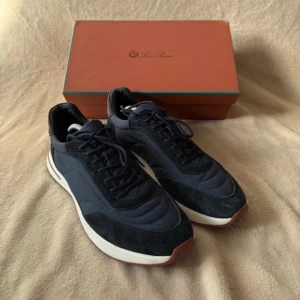 Mörkblå sneakers från Loro Piana - Snygga mörkblå sneakers från Loro Piana med ovandel i mocka och textil, vita sulor och bruna detaljer undertill. Skorna har klassisk snörning och en stilren, sportig design som passar perfekt till jeans eller chinos.