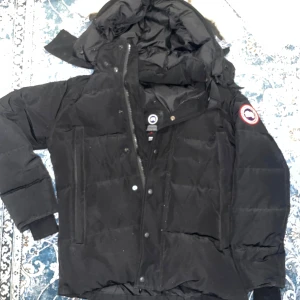 Svart dunjacka från Canada Goose XS - Säljer en svart dunjacka från Canada Goose i storlek XS. Jackan har huva, dragkedja och knappar framtill samt klassisk logga på ärmen. Fodrad med dun och fjädrar för extra värme, perfekt för kalla dagar. Snygg och tidlös design med flera praktiska fickor.