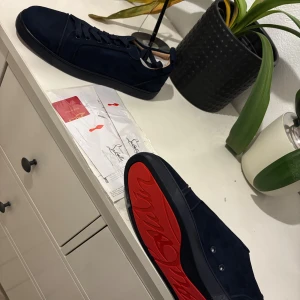 Mörkblå Christian Louboutin sneakers - Säljer ett par mörkblå sneakers från Christian Louboutin med klassisk röd sula och snygg snörning. Skorna är i mocka och har en stilren, låg profil som passar perfekt till streetwear eller en mer uppklädd look.