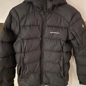 Svart Peak Performance pufferjacka S - Svart pufferjacka från Peak Performance i storlek S. Jackan har huva, dragkedja framtill och två sidofickor. Diskret logga på bröstet och ärmen. Perfekt för kalla dagar med sin fluffiga och isolerande design. Materialet är syntet och dun. Ett litet hål vid ena ärmen men annars i bra skick (kolla sista bilden)