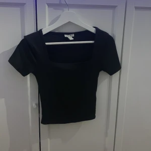 Svart croppad topp från Amisu - Svart croppad topp från Amisu med fyrkantig halsringning och korta ärmar. Enkel och stilren design som är lätt att matcha med olika outfits. Mjuk och stretchig polyester som sitter snyggt och bekvämt. Perfekt för dig som gillar minimalistisk stil.