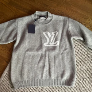 Grå stickad tröja Louis Vuitton - Snygg grå stickad tröja från Louis Vuitton med stort vitt LV-logo framtill. Tröjan har ribbad krage, ärmslut och nederkant. Perfekt för dig som vill ha en exklusiv och stilren look med lyxig känsla.