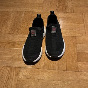 Svarta slip-on sneakers med vit sula från Zara som man kan ha som grmpaskor på idrotten. - Svarta slip-on sneakers från Salomon med stickad ovandel och vit platt sula. Skorna har en rund tå och en diskret röd detalj på plösen. Praktisk dragflik bak gör dem enkla att ta på. Perfekt för dig som gillar stilren och sportig look.